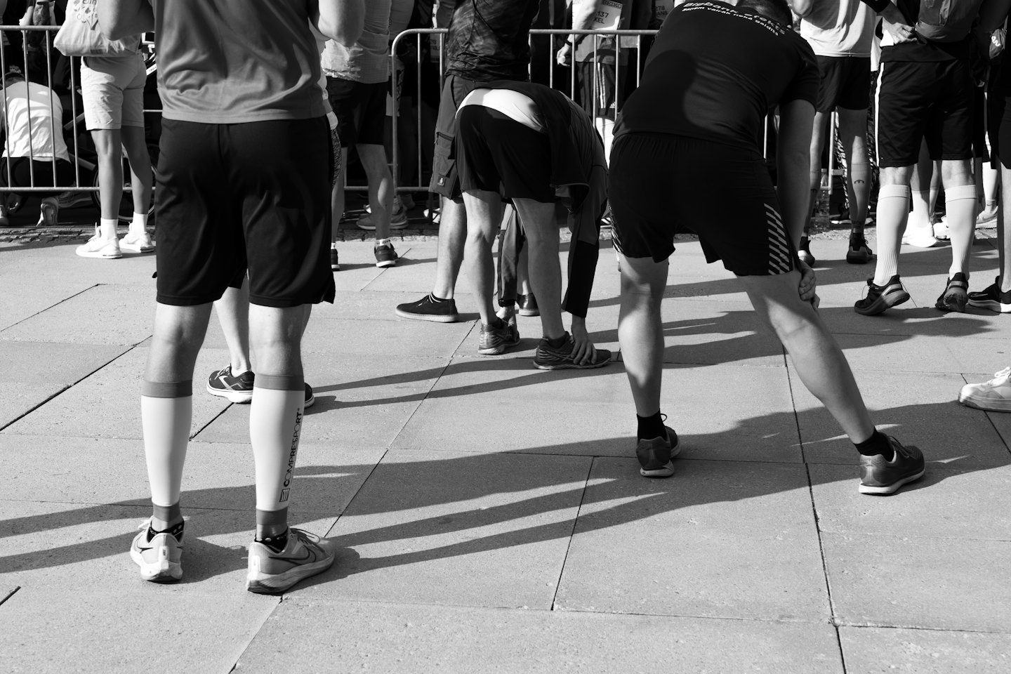 Runners stretching their legs on pavement near a barrier.